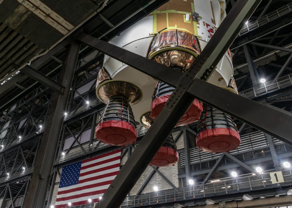 The bottom of the SLS core stage with four engines under red caps, lifted above the transfer aisle of the Vehicle Assembly Building