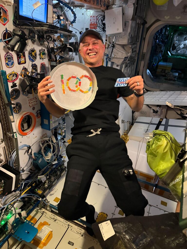 NASA astronaut Anil Menon holds a patch and a tray of snacks marking his 100th day in space aboard the space station