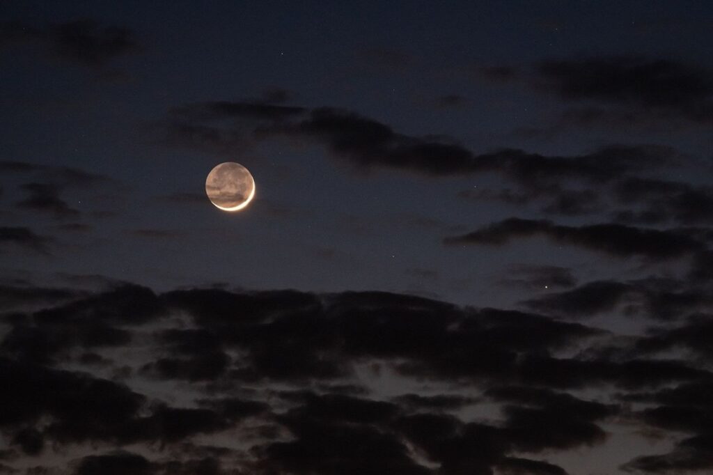 A crescent Moon in a dark, cloudy sky with the rest of its near side dimly visible