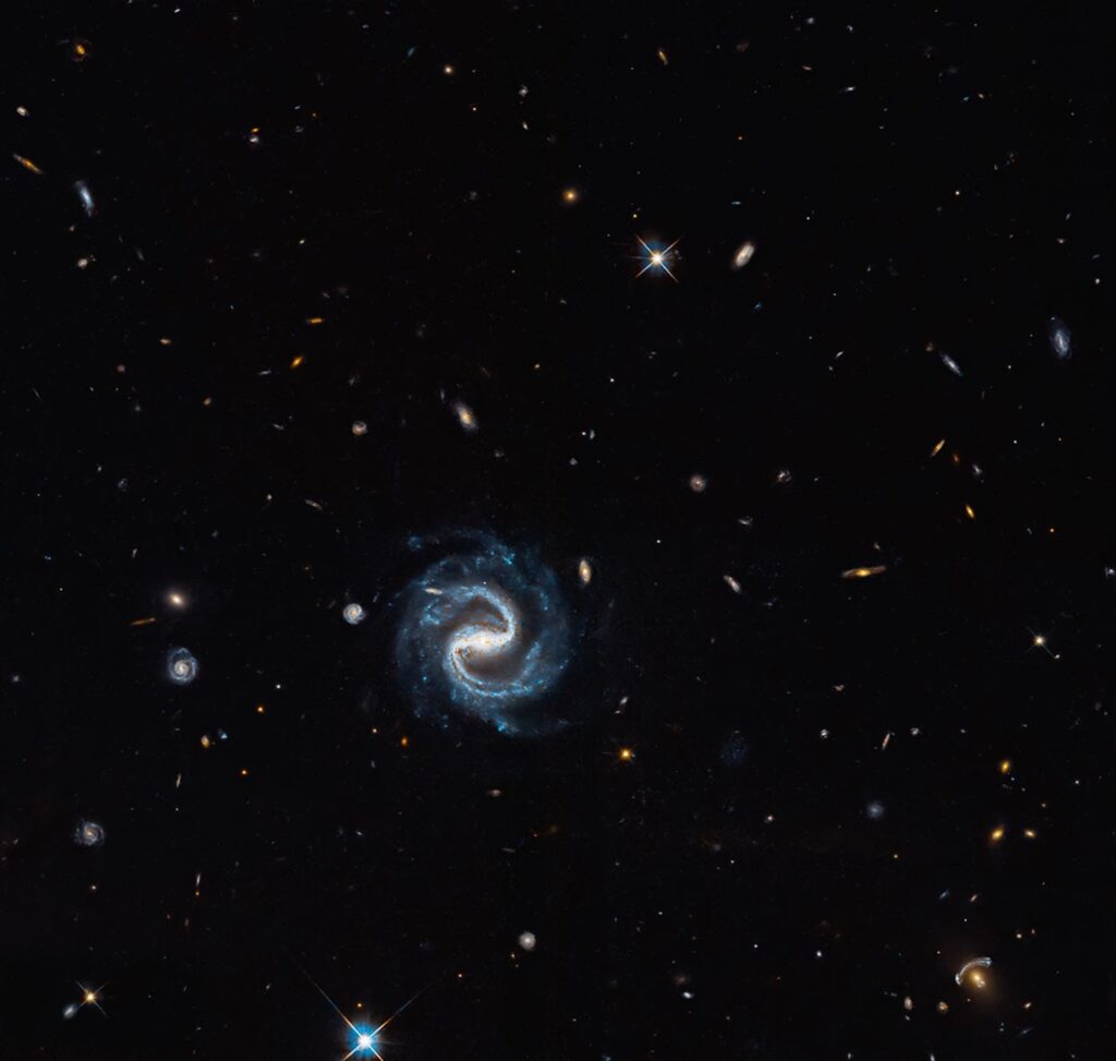 A spiral galaxy with two distinct arms in the foreground against many distant galaxies, with a gravitational lens arc in the lower right, seen by Hubble