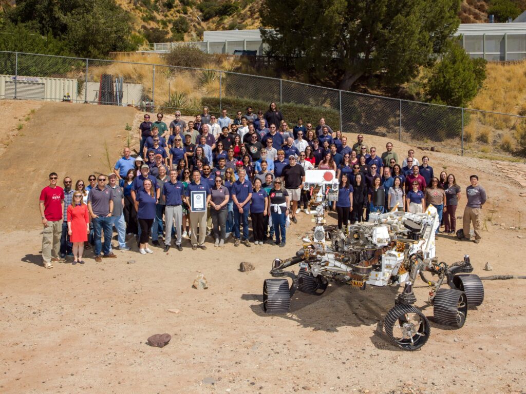 About 100 members of the Perseverance team beside the full-scale OPTIMISM rover model in the Mars Yard at JPL, holding a Guinness World Records certificate