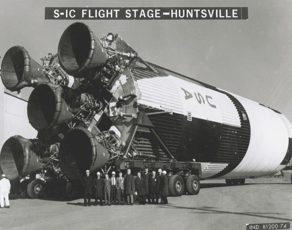 A small group of officials dwarfed by the Saturn V first stage at Marshall in Huntsville, Alabama